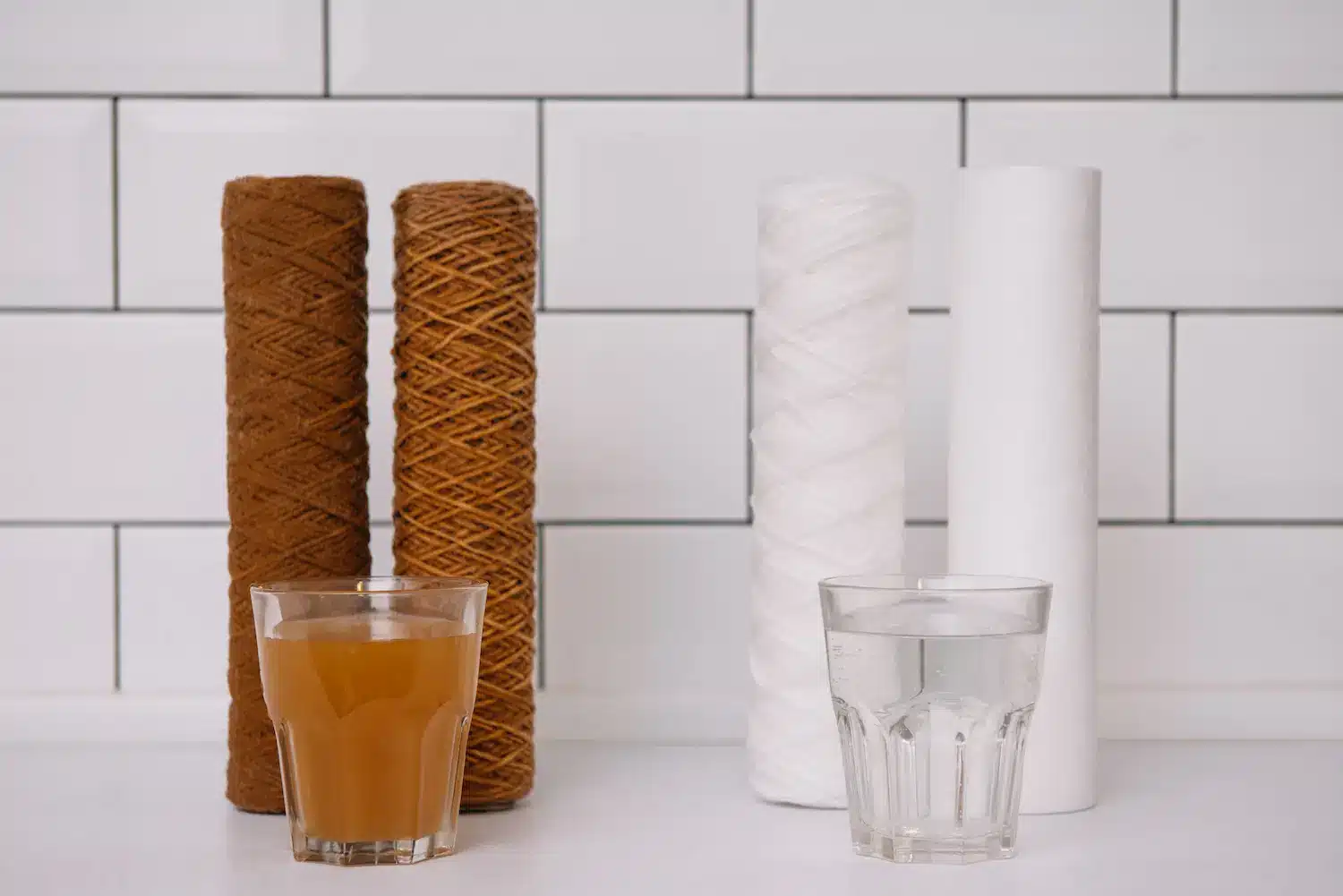 Studio comparison of iron fouled and clean water filter cartridges beside glasses of water