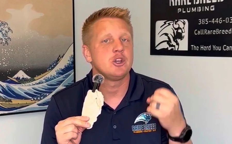 Court Lundberg on camera holding a scaled water heater component, with an older Rare Breed sign showing a retired phone number behind him