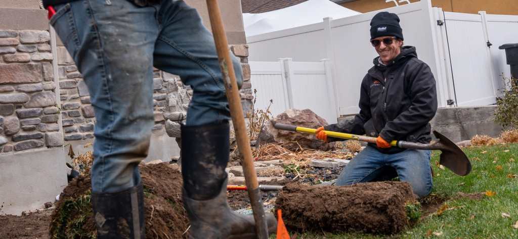 Two technicians hand-digging a service line trench beside a Utah home